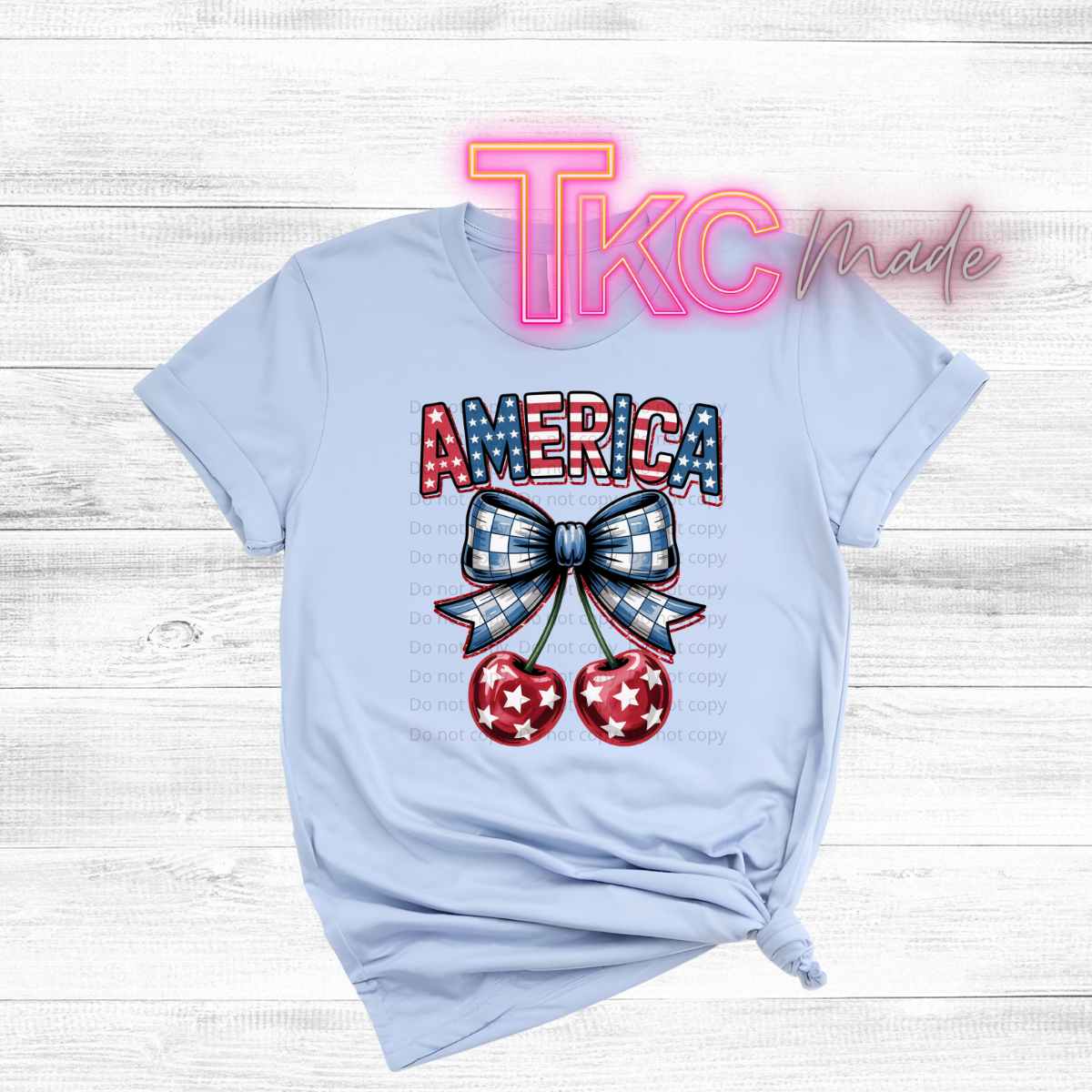 Light blue t-shirt with 'AMERICA' and bow design on a wooden surface, featuring 'TKC Made' branding.