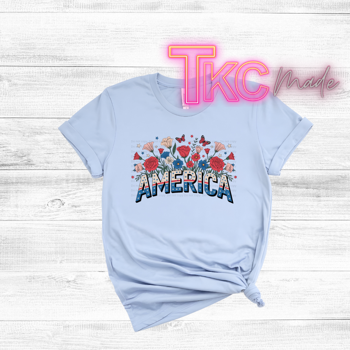 Light blue t-shirt with 'America' and floral design on a wooden surface, featuring 'TKC Made' text.