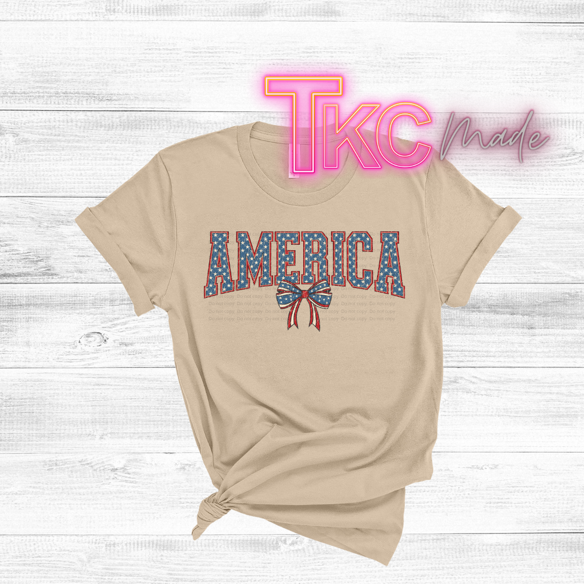 Beige t-shirt with 'AMERICA' print on a wooden surface, featuring 'TKC Made' logo.