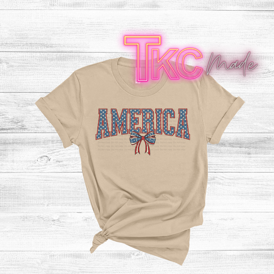 Beige t-shirt with 'AMERICA' print on a wooden surface, featuring 'TKC Made' logo.