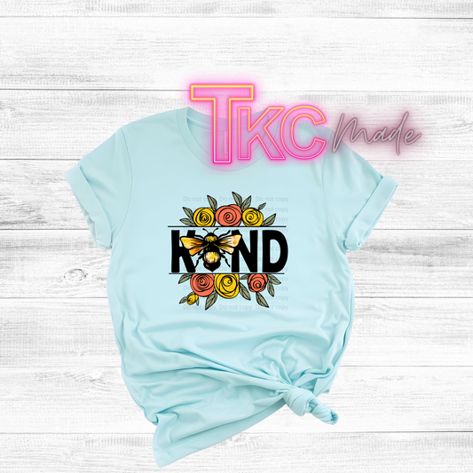 Light blue t-shirt with a bee and flower design on a wooden surface with 'TKC Made' branding.