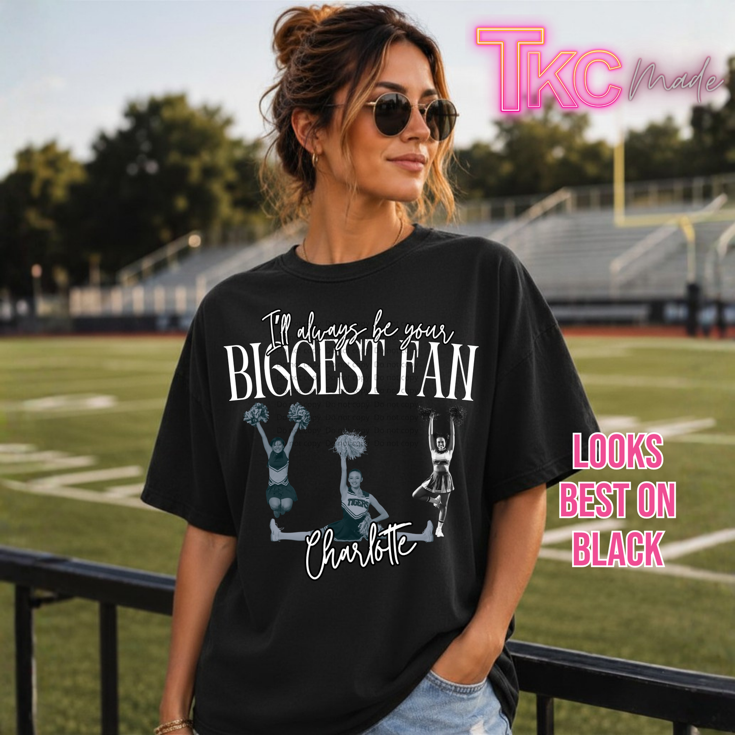 A woman wearing a black graphic t-shirt with white text reading 'I'll always be your BIGGEST FAN Charlotte' and cheerleader illustrations, standing on a football field. Pink text on the right says 'LOOKS BEST ON BLACK' and a watermark says 'TKC Made'.