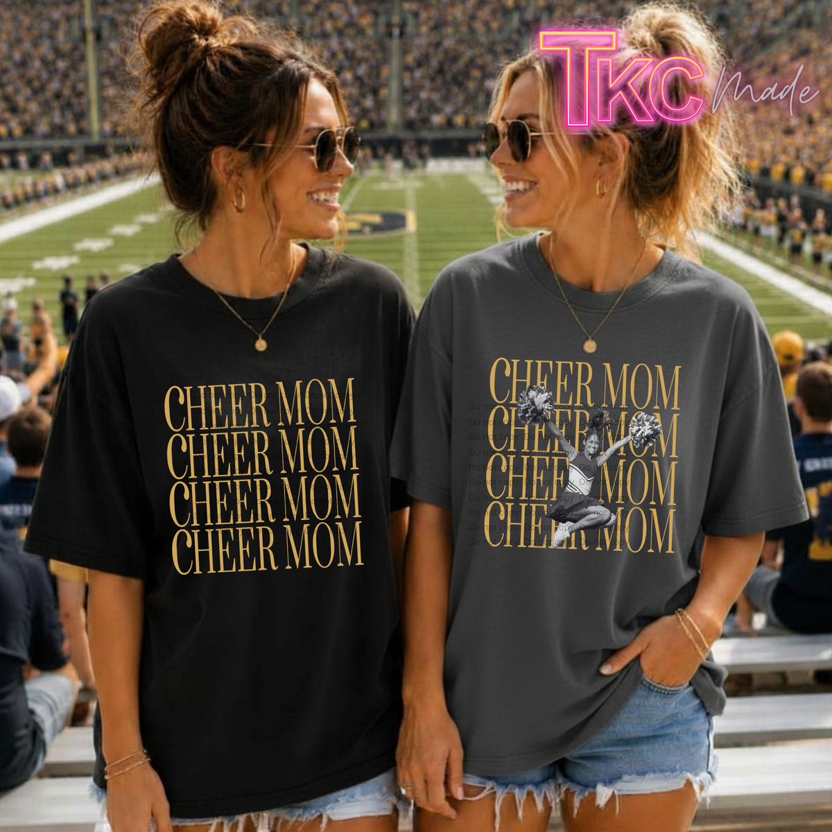 Two women standing in a stadium wearing oversized graphic t-shirts. The shirt on the left is black with gold 'CHEER MOM' text, and the shirt on the right is dark gray with gold 'CHEER MOM' text and a black graphic of a cheerleader.