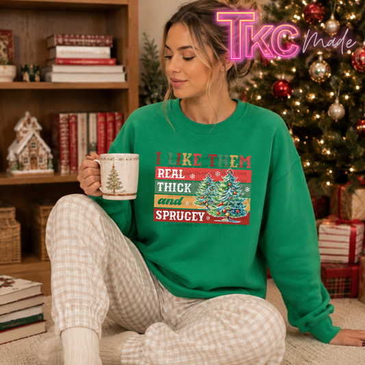 A woman sitting on a carpet in front of a decorated Christmas tree, wearing a bright green crewneck sweatshirt with a colorful holiday graphic and text, paired with beige plaid pajama pants. She is holding a white ceramic mug.