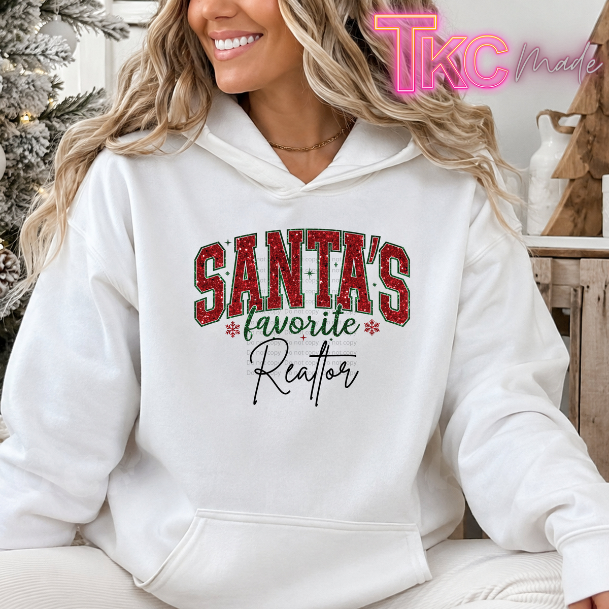 A woman wearing a white pullover hoodie with a festive graphic that reads 'SANTA'S favorite Reator' in red, green, and black lettering. The photo is taken in a cozy indoor setting with a Christmas tree in the background, and a pink 'TKC Made' watermark is visible in the top right corner.