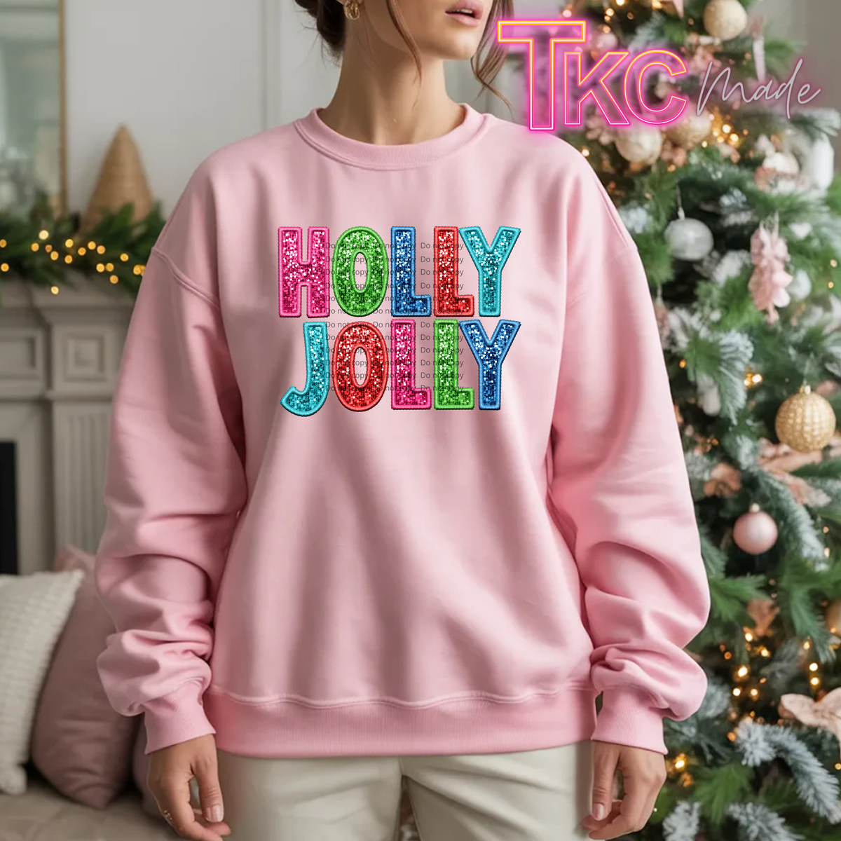 A woman wearing a light pink crewneck sweatshirt with 'HOLLY JOLLY' printed in colorful glitter-texture letters, posed in front of a decorated Christmas tree. A 'TKC Made' watermark is visible in the top right corner.