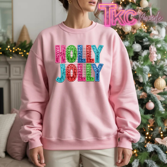 A woman wearing a light pink crewneck sweatshirt with 'HOLLY JOLLY' printed in colorful glitter-texture letters, posed in front of a decorated Christmas tree. A 'TKC Made' watermark is visible in the top right corner.