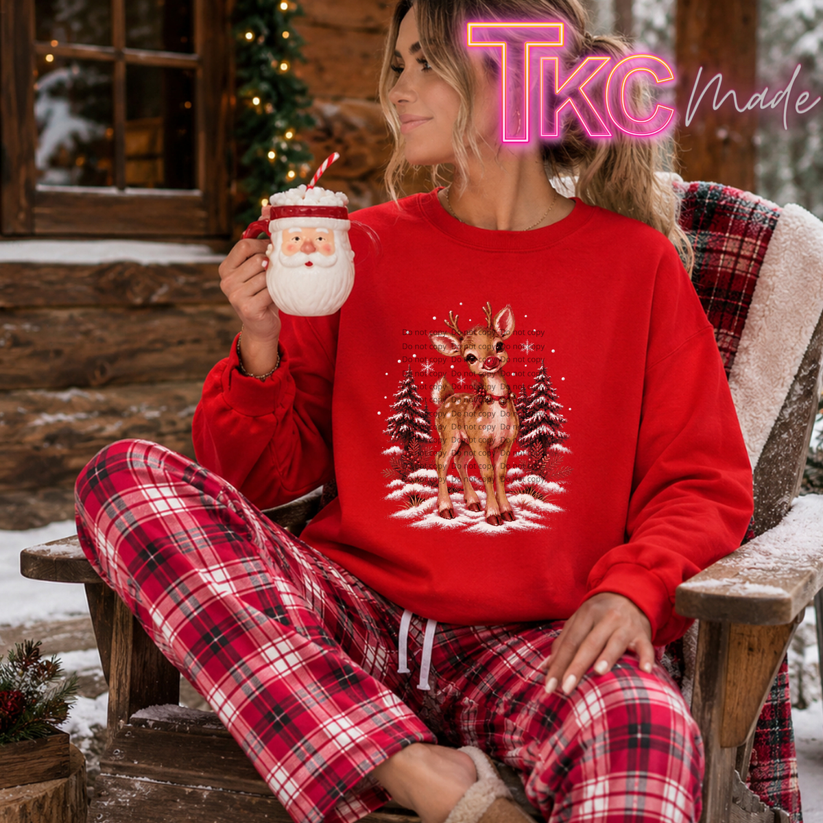 A woman sitting in a wooden chair outdoors wearing a bright red Christmas sweater with a reindeer graphic and matching red-and-black plaid pajama pants. She is holding a Santa-themed mug, with a rustic cabin and snowy setting in the background.