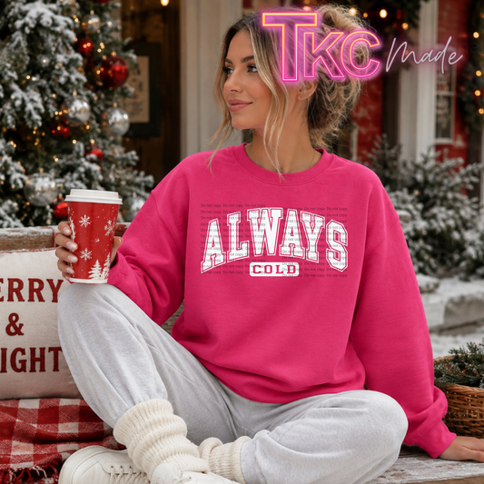 A woman sits cross-legged outdoors in a cozy holiday setting, wearing a bright pink crewneck sweatshirt with 'ALWAYS COLD' printed in white block letters across the chest. She holds a red holiday coffee cup.