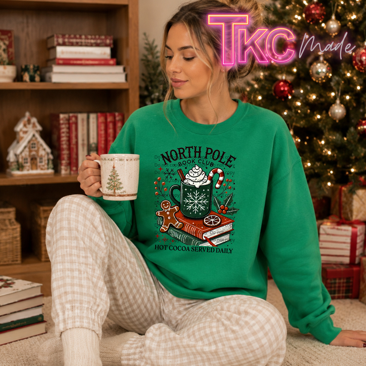 A woman sitting on a carpet in a cozy room, wearing a bright green crewneck sweatshirt with a 'North Pole' hot cocoa graphic. She is holding a mug and wearing plaid pajama pants, with a decorated Christmas tree and bookshelves in the background.