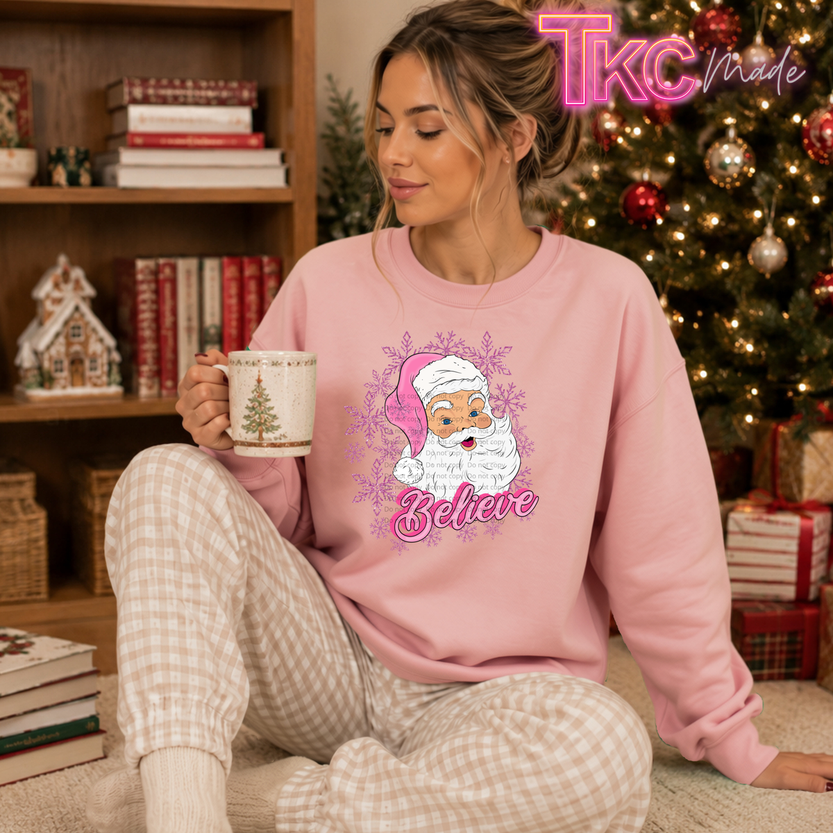 A woman sits on the floor in a cozy living room setting, wearing a pink crewneck sweatshirt with a festive graphic of Santa Claus and the word 'Believe' in pink script. She is holding a white mug and wearing beige and white plaid pajama pants.