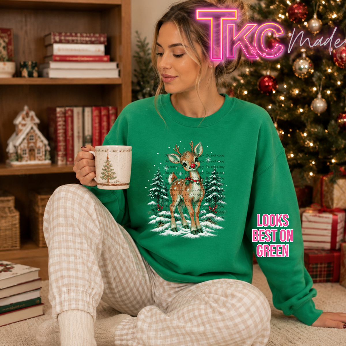 A woman sits on a carpet in a cozy holiday room wearing a bright green crewneck sweatshirt with a festive Christmas graphic of a reindeer in the snow. She holds a mug, with a bookshelf and a decorated Christmas tree in the background.