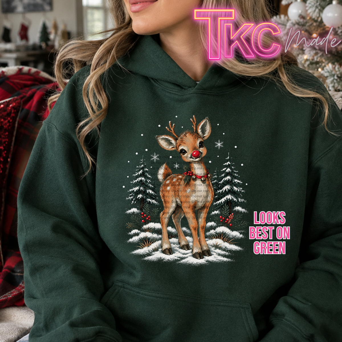 A woman wearing a dark forest green hoodie with a Christmas graphic of a plaid deer in a snowy landscape. The hoodie has a pink 'TKC made' watermark overlay and the text 'LOOKS BEST ON GREEN' on the right side.
