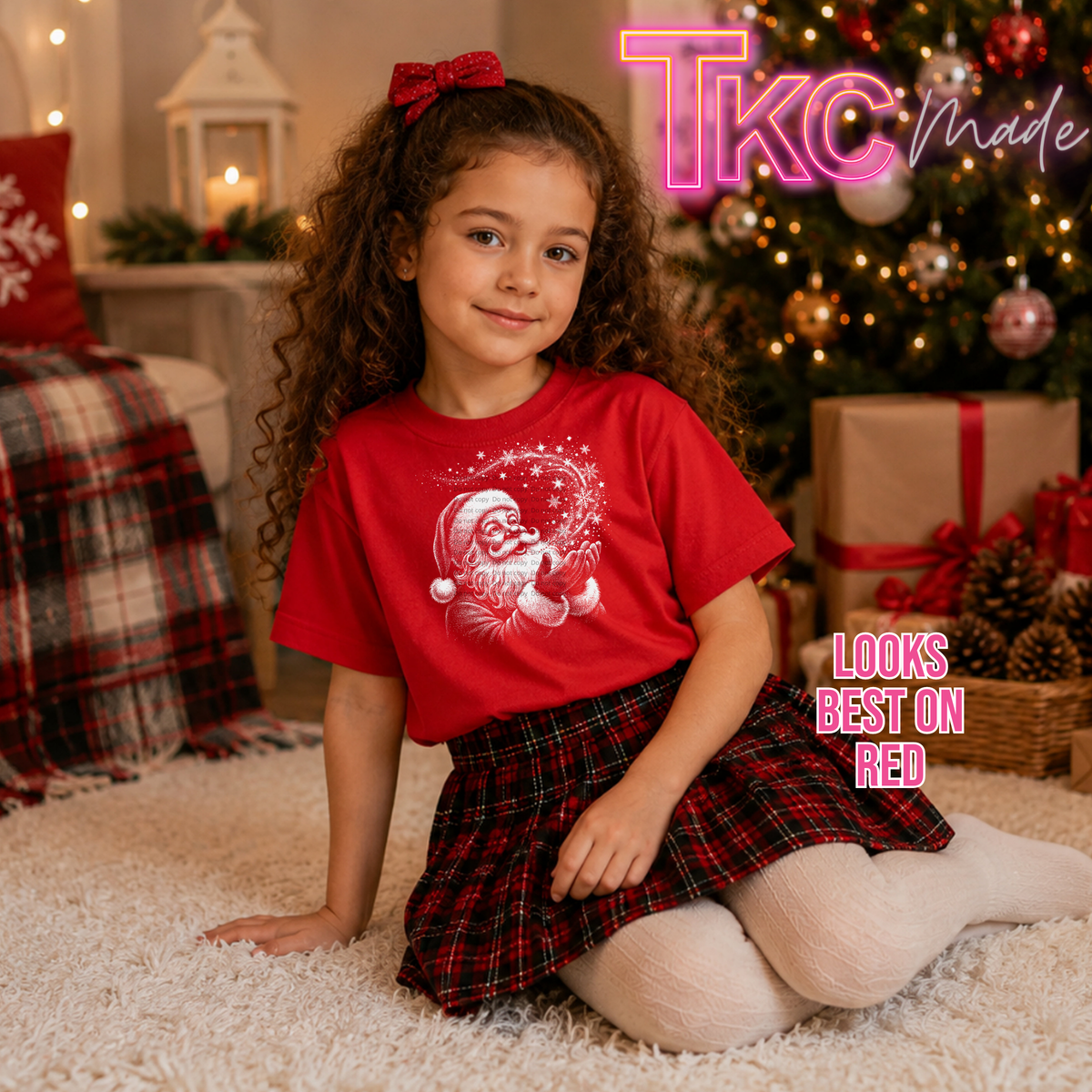 A young girl with curly brown hair sits on a white shag rug in a cozy Christmas-themed room, wearing a red short-sleeve graphic T-shirt with a white Santa Claus snow globe design and a red plaid skirt. The image includes 'TKC Made' and 'LOOKS BEST ON RED' text overlays.