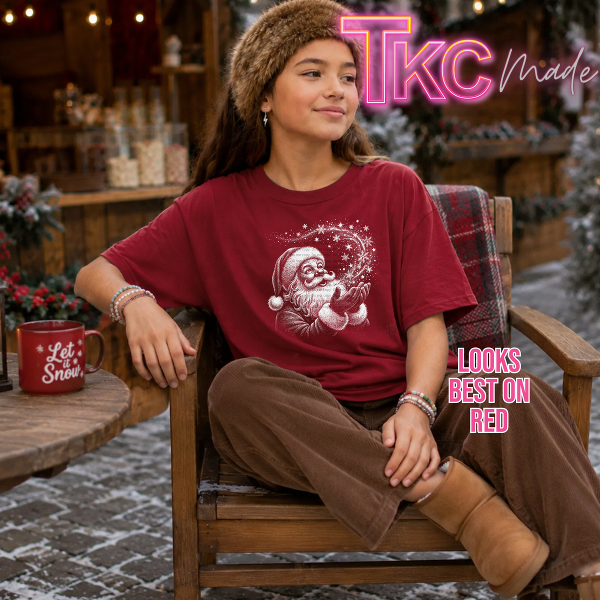 A young woman sits in a wooden chair wearing a festive red graphic t-shirt with a white Santa Claus design, brown pants, and a brown fur hat. The background depicts a cozy, snowy holiday village scene with a wooden table holding a red mug that says 'Let Snow Snow'.
