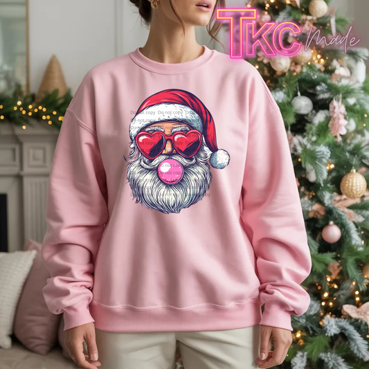 A woman wearing a light pink crewneck sweatshirt with a graphic of a Santa Claus face wearing red heart-shaped sunglasses and blowing a pink bubblegum bubble. The background shows a decorated Christmas tree and a fireplace.