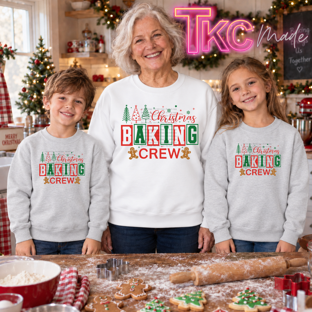 Three people standing together in a decorated holiday kitchen wearing matching light gray crewneck sweatshirts with a 'Christmas Baking Crew' graphic. The image has a pink 'TKC made' watermark overlay in the top right corner.