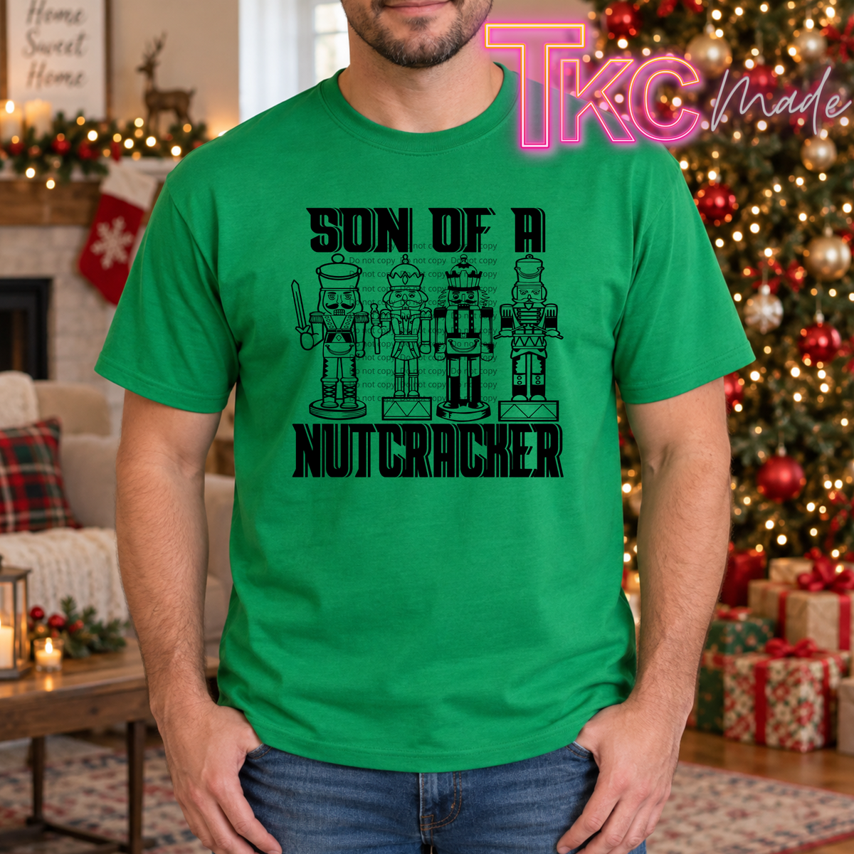 A man wearing a bright green t-shirt with a black graphic that reads 'SON OF A NUTCRACKER' above four nutcracker figures. The photo is taken in a cozy, decorated Christmas living room with a fireplace, stockings, and a lit tree in the background.
