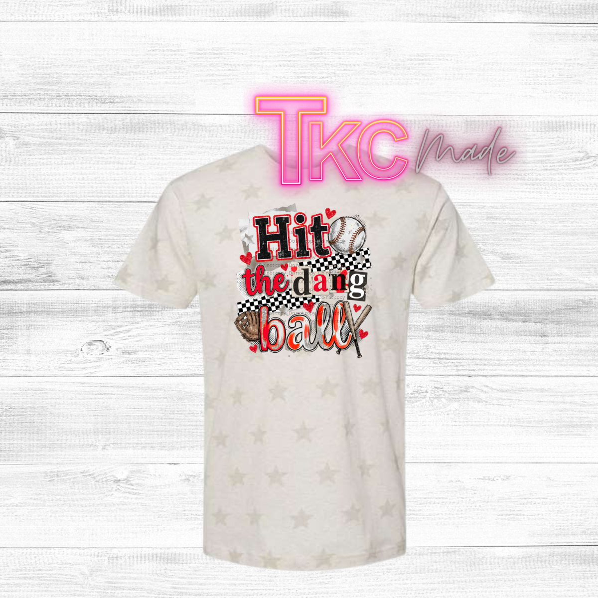 White t-shirt with sports-themed design "hit the dang ball" and 'TKC made' text on a light wooden background