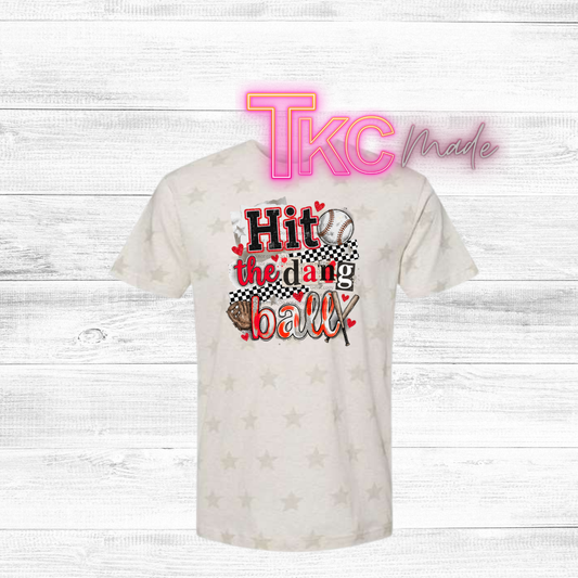 White t-shirt with sports-themed design "hit the dang ball" and 'TKC made' text on a light wooden background