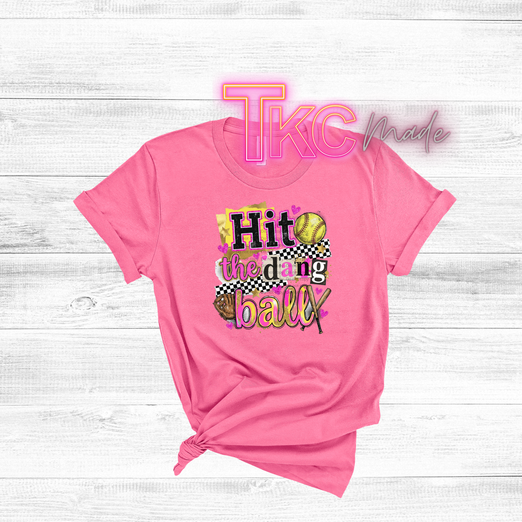 Pink t-shirt with sports-themed design "hit the dang ball" and 'TKC made' text on a light wooden background