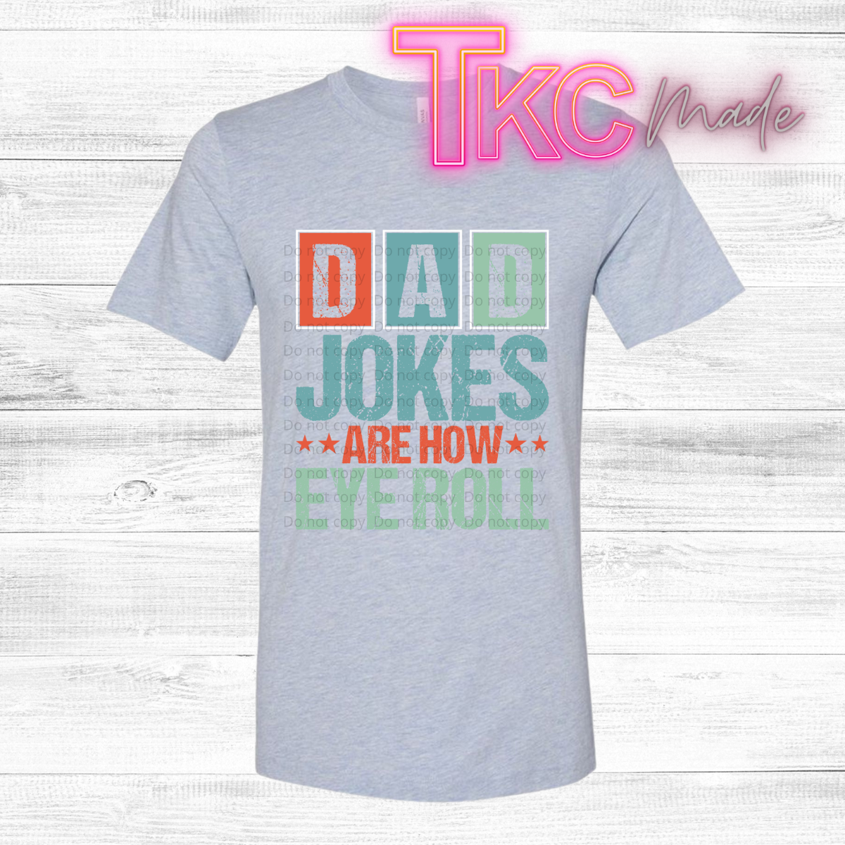 Gray t-shirt with colorful text 'Dad jokes are how eye roll' on a wooden background with 'TKC Made' branding.