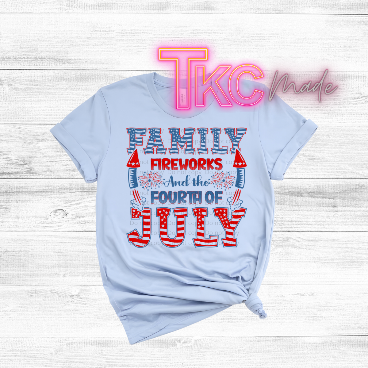 Light blue t-shirt with 'Family Fireworks and the Fourth of July' text on a wooden surface, featuring the brand 'TKC Made'.