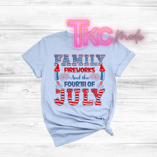 Light blue t-shirt with 'Family Fireworks and the Fourth of July' text on a wooden surface, featuring the brand 'TKC Made'.