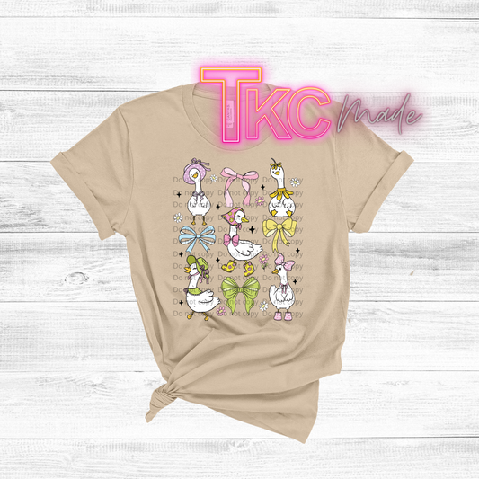 Beige t-shirt with cartoon ducks and bows on a wooden surface, featuring 'TKC Made' text.