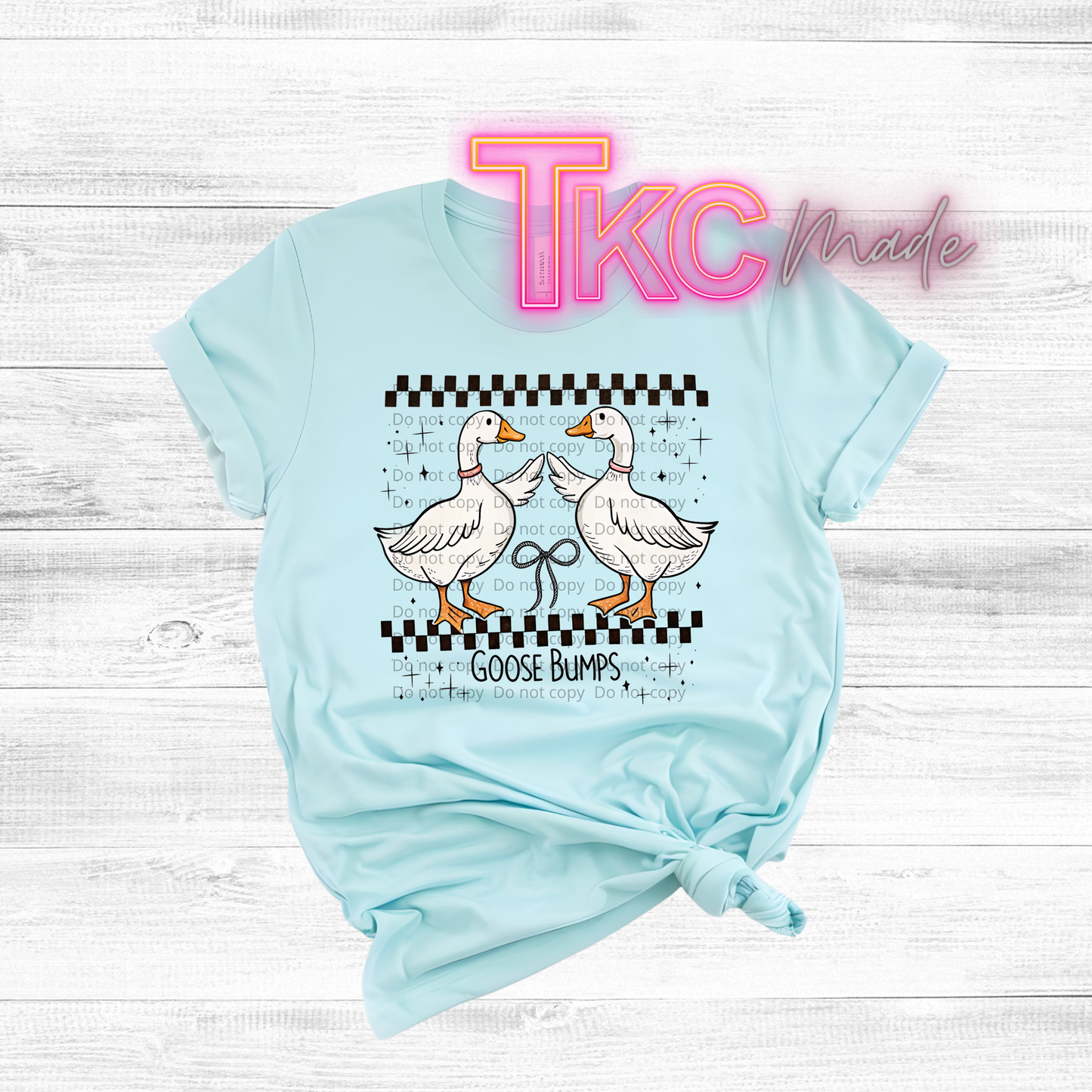Light blue t-shirt with cartoon geese and 'TKC made' text on a wooden surface