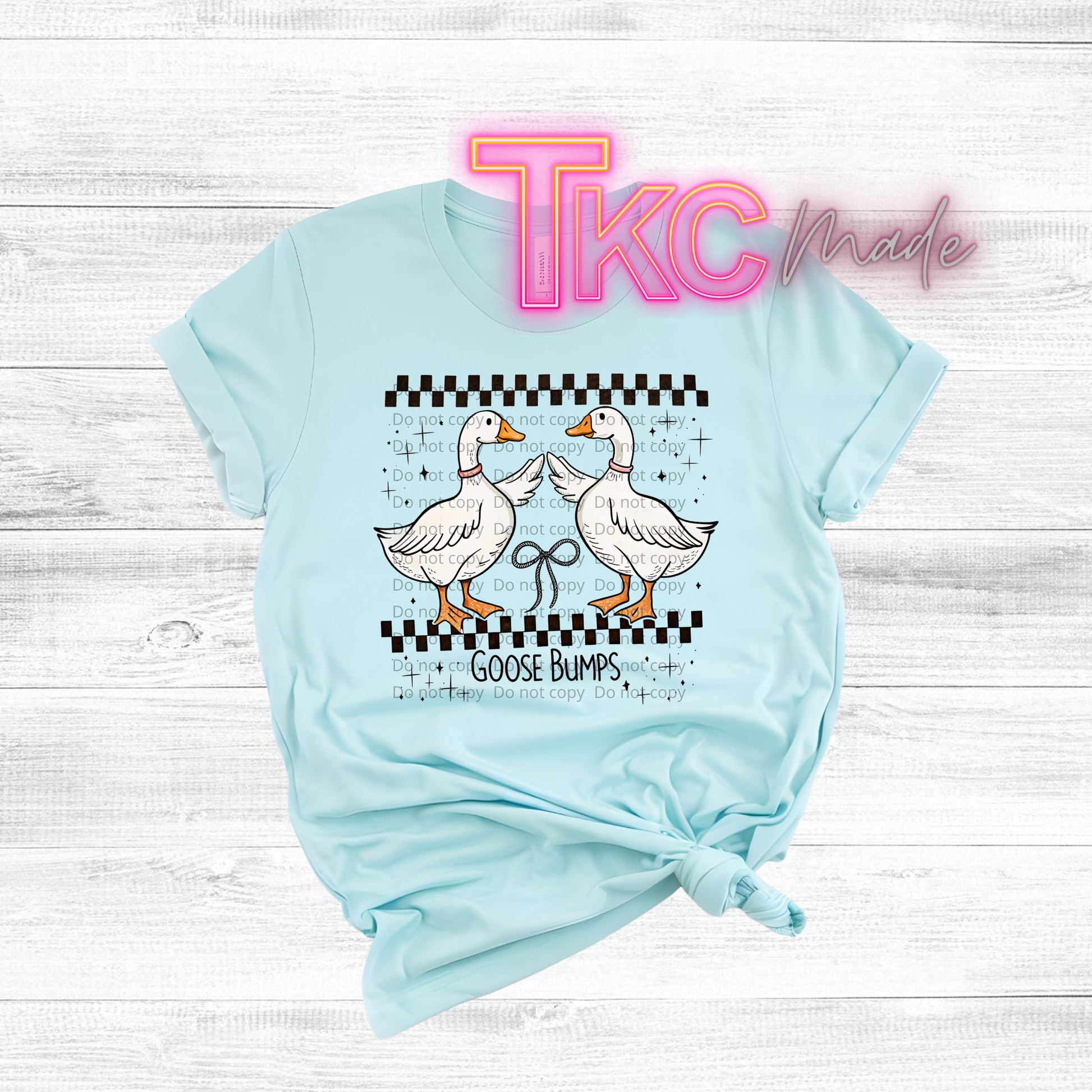 Light blue t-shirt with cartoon geese and 'TKC made' text on a wooden surface