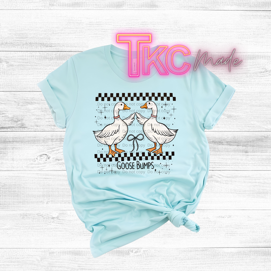 Light blue t-shirt with cartoon geese and 'TKC made' text on a wooden surface