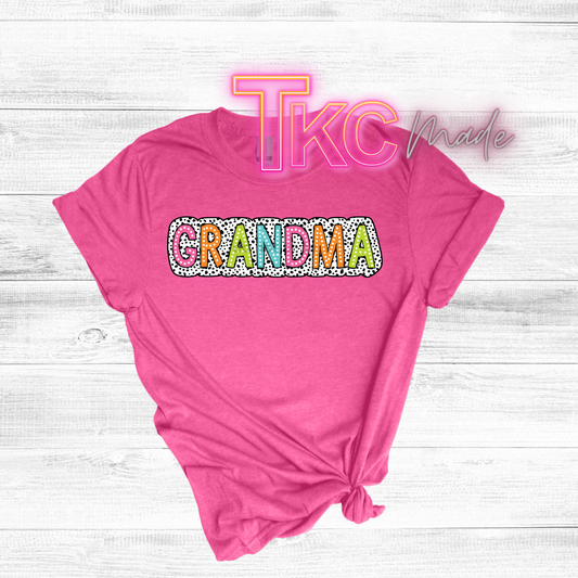Pink t-shirt with 'GRANDMA' text on a wooden surface, featuring 'TKC Made' branding.