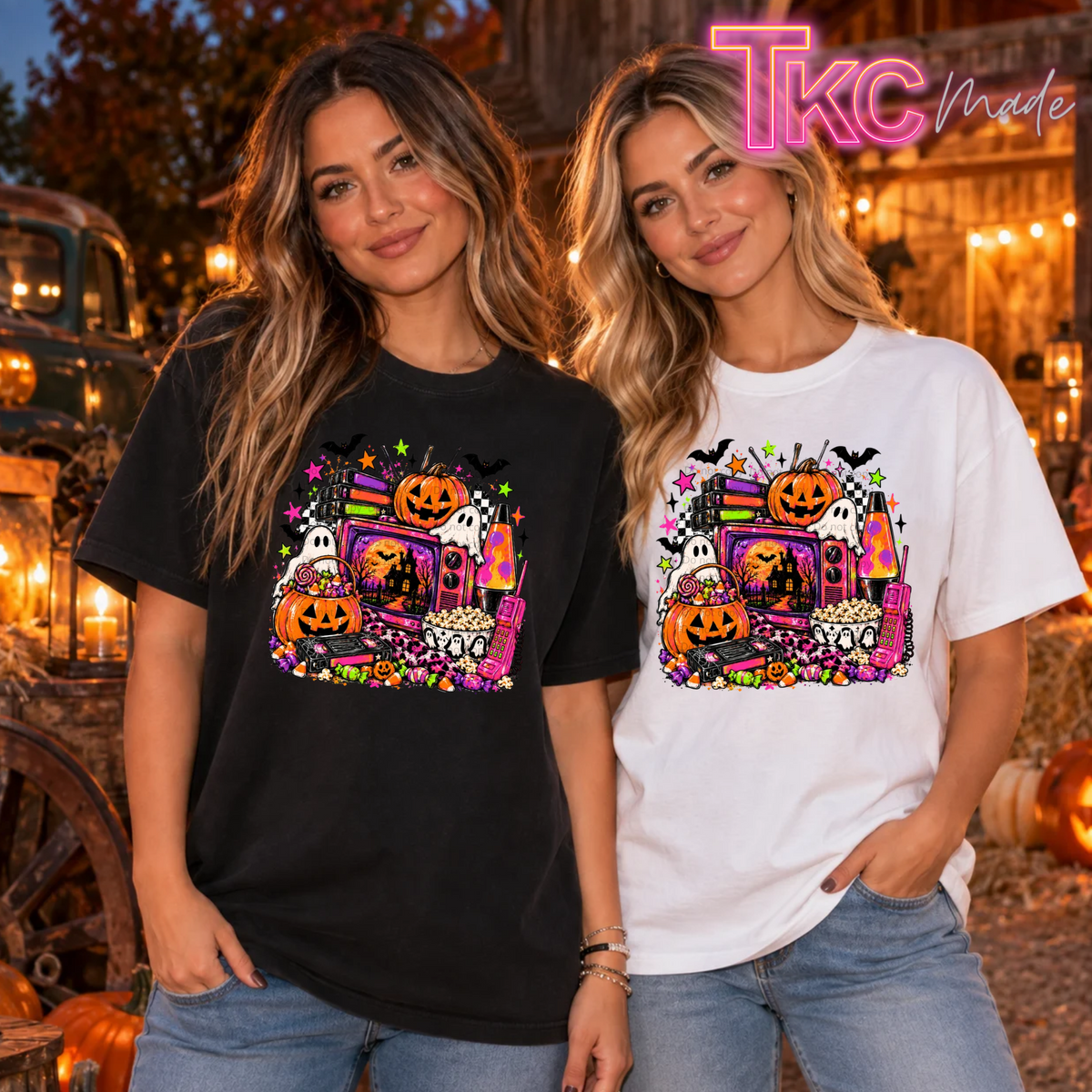 Two women wearing matching Halloween graphic t-shirts in a rustic outdoor setting at night. The woman on the left wears a black t-shirt and the woman on the right wears a white t-shirt, both featuring a colorful graphic of a haunted truck with pumpkins, ghosts, bats, and candy corn like a 90's Halloween theme.