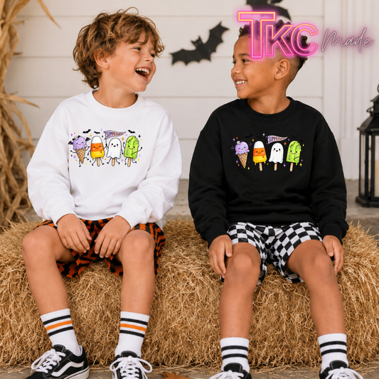Two children sitting on hay bales wearing matching graphic sweatshirts. The child on the left wears a white sweatshirt with a colorful ice cream cone and ghost Halloween graphic, paired with orange-and-black checkered shorts.