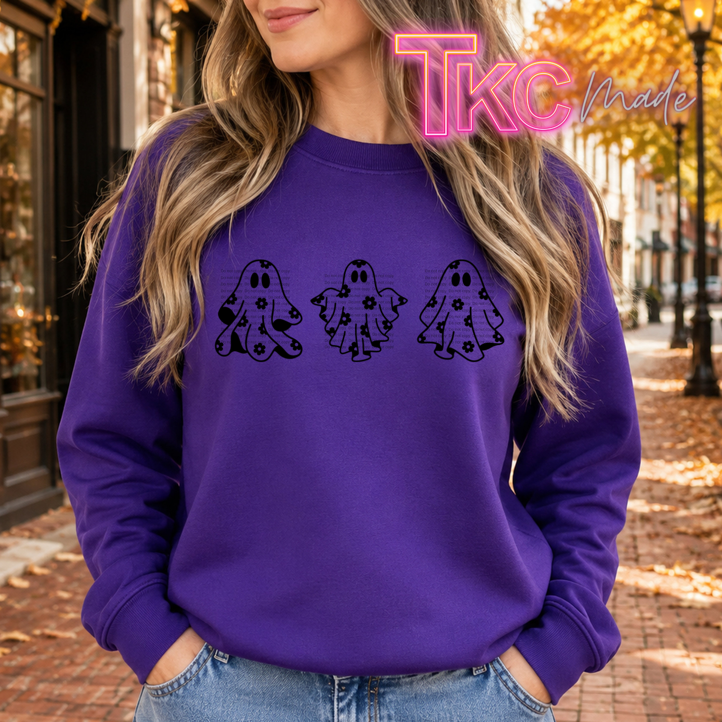 A woman wearing a purple crewneck sweatshirt with three black ghost graphics across the chest, set against an autumn street background. A pink 'TKC Made' watermark is visible in the top right.