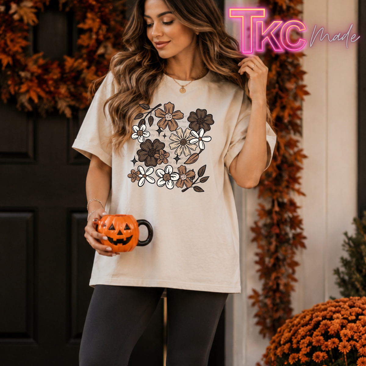 A woman wearing a cream-colored t-shirt with a brown and white floral graphic, holding a small orange jack-o'-lantern mug, posed in front of a decorated autumn door.