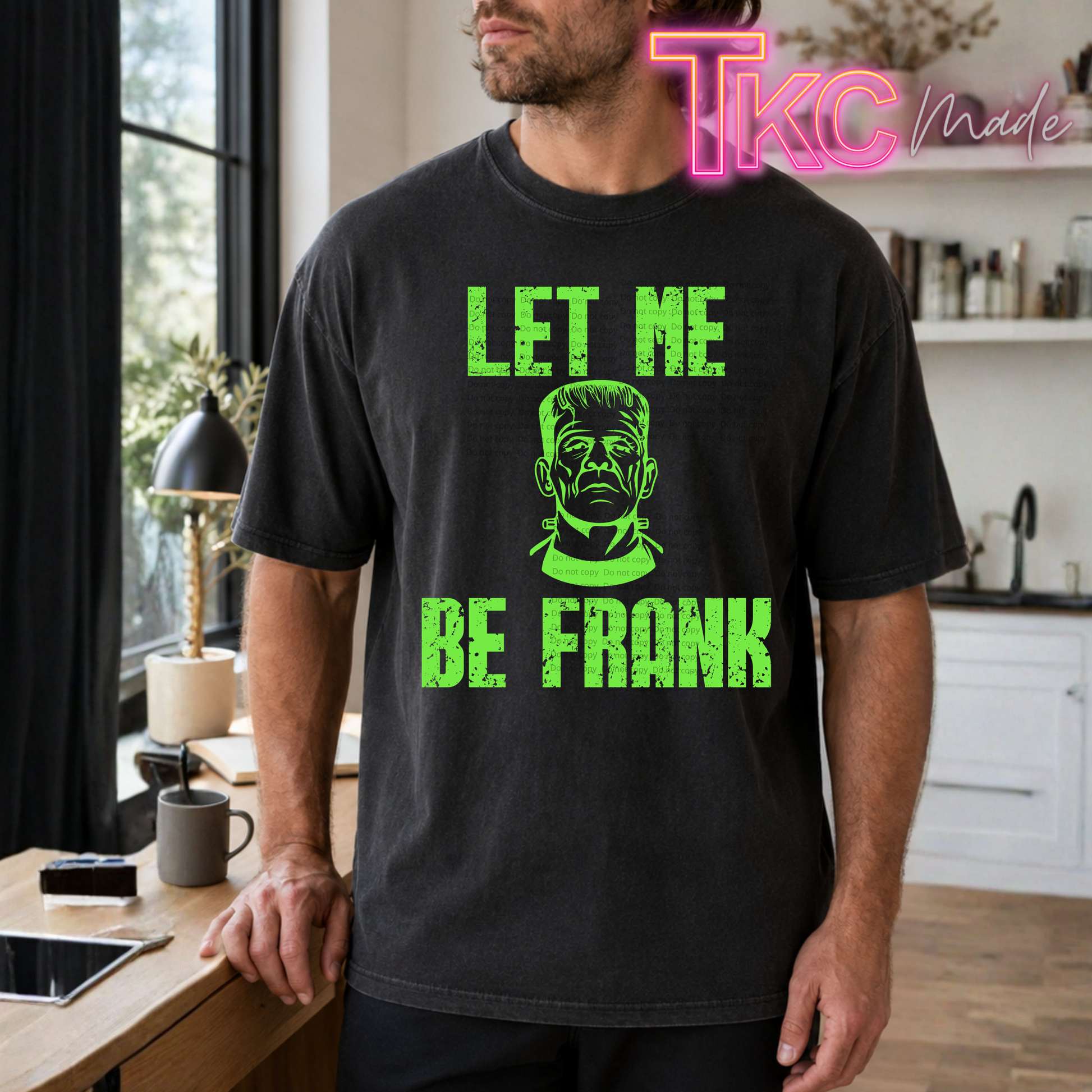 A man wearing a black graphic t-shirt with a bright green distressed print that reads 'LET ME BE FRANK' and features a Frankenstein's monster face illustration. The photo is taken in a modern kitchen setting, with a 'TKC Made' watermark in the top right corner.