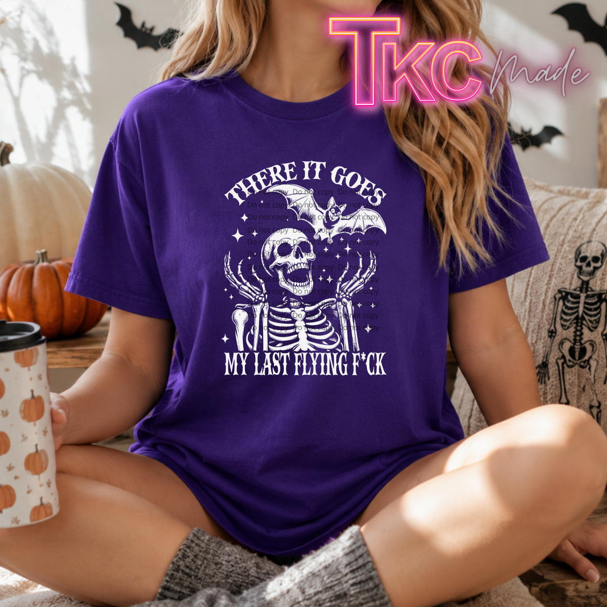 A woman wearing a purple short-sleeve graphic T-shirt with a white skeleton and bat design and the text 'THERE IT GOES MY LAST FLYING F*CK'. She is sitting in a cozy indoor setting with Halloween decorations, holding a pumpkin-patterned mug.