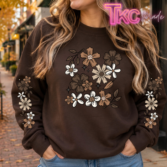 A woman wearing a dark brown crewneck sweatshirt with a floral graphic on the chest and sleeves. The sweatshirt features white, tan, and brown flowers with black outlines.