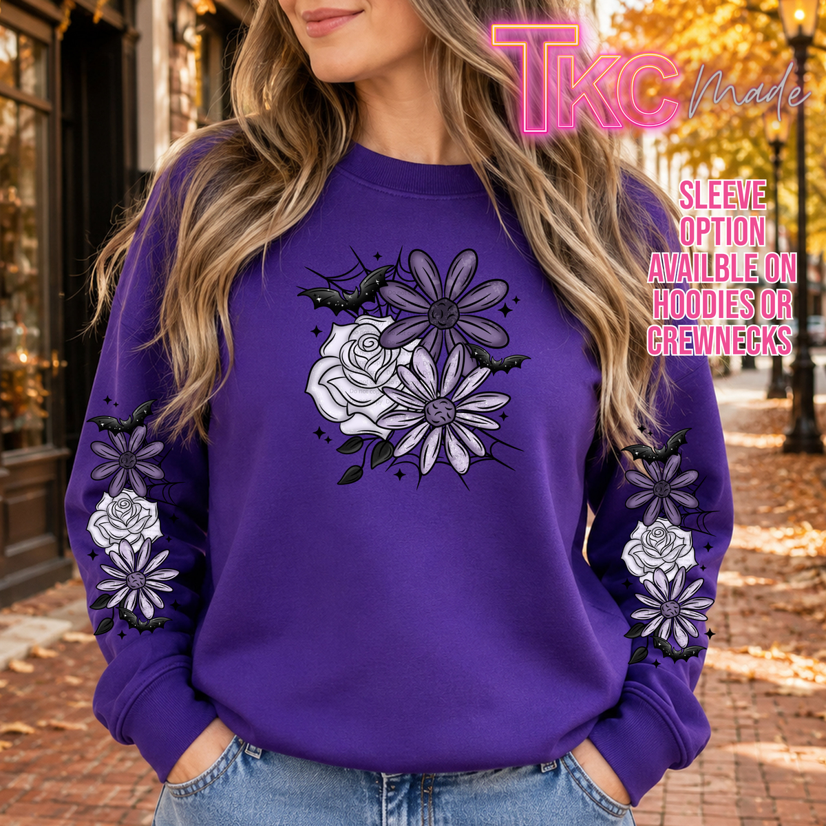 A woman wearing a purple sweatshirt with a graphic of white and purple roses, black bats, and purple daisies on the front, with matching graphics on the sleeves. The background is an outdoor street scene with autumn leaves.