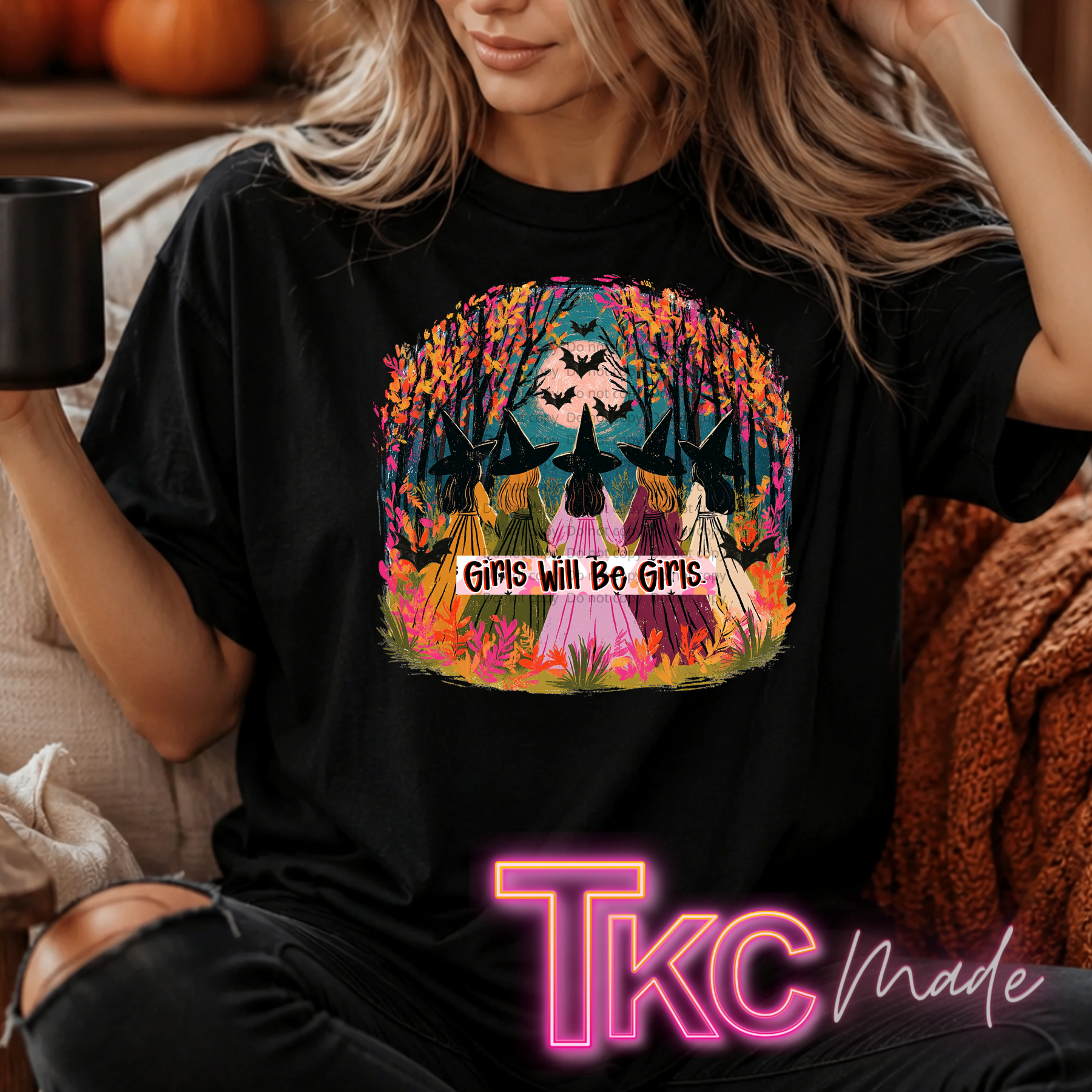A woman wearing a black graphic T-shirt with a colorful Halloween-themed design showing witches in hats under autumn trees and the text 'Girls Will Be Girls.' She is seated indoors holding a black mug, with a cozy blanket and pumpkins in the background.