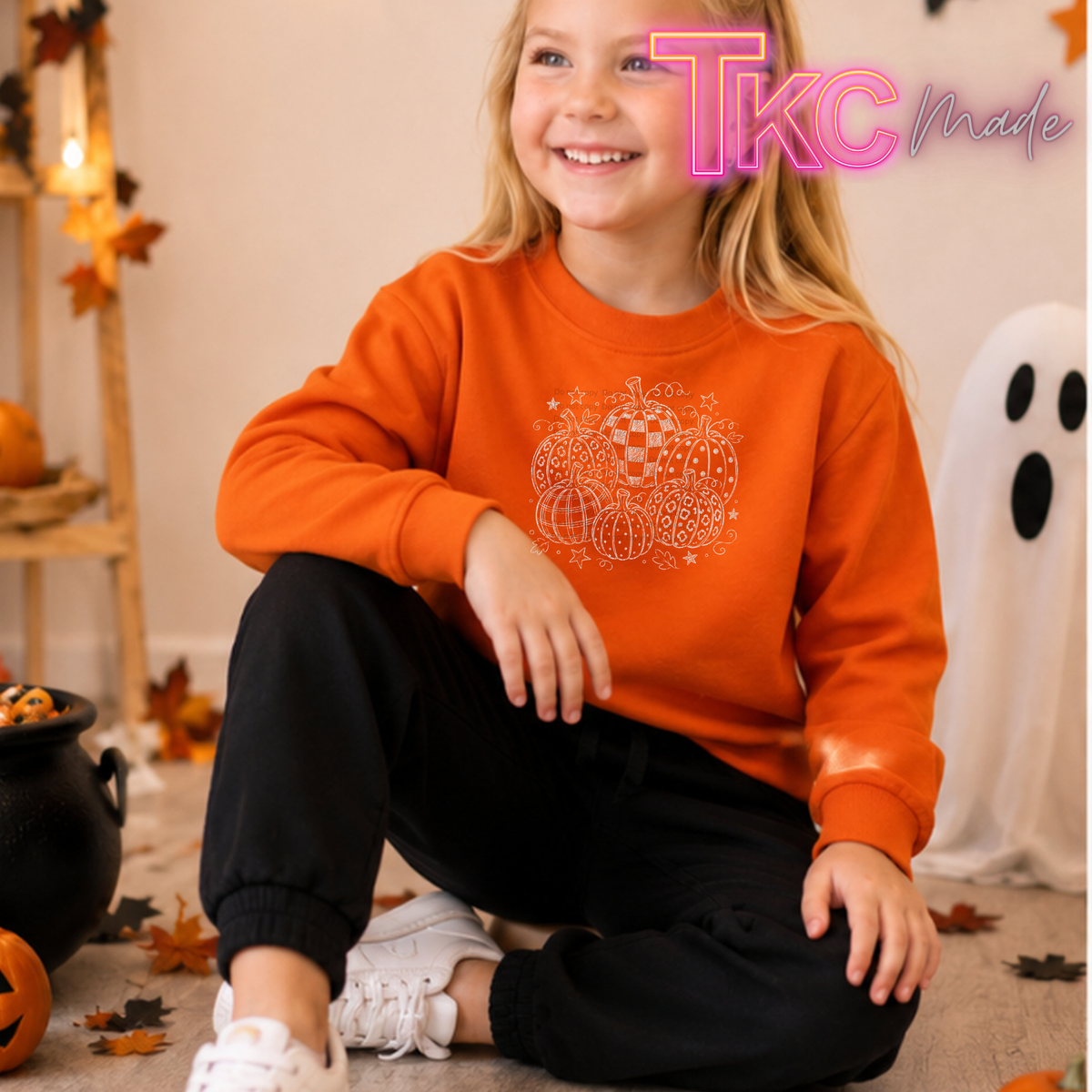 A young girl sitting on a carpeted floor wearing an orange sweatshirt with a white pumpkin graphic and black pants, surrounded by Halloween decorations including a ghost, pumpkins, and a cauldron. A 'TKC Made' watermark is visible in the top right corner.