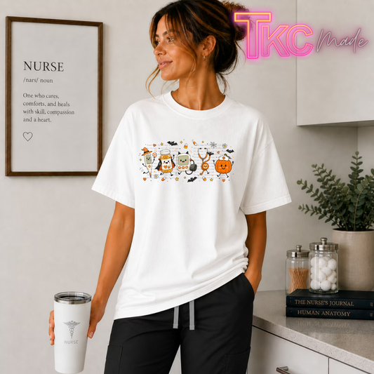 A woman wearing a white crewneck t-shirt with a colorful cartoon graphic of cute faces on a pumpkin, pill bottle, thermometer and stethoscope dressed as Halloween characters, standing in a doctor office setting. She is holding a white travel tumbler with a medical cross logo.