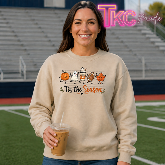 A woman stands on a football field wearing a beige crewneck sweatshirt with a colorful graphic of autumn sports items and the text 'Tis the Season'. She is holding an iced coffee.