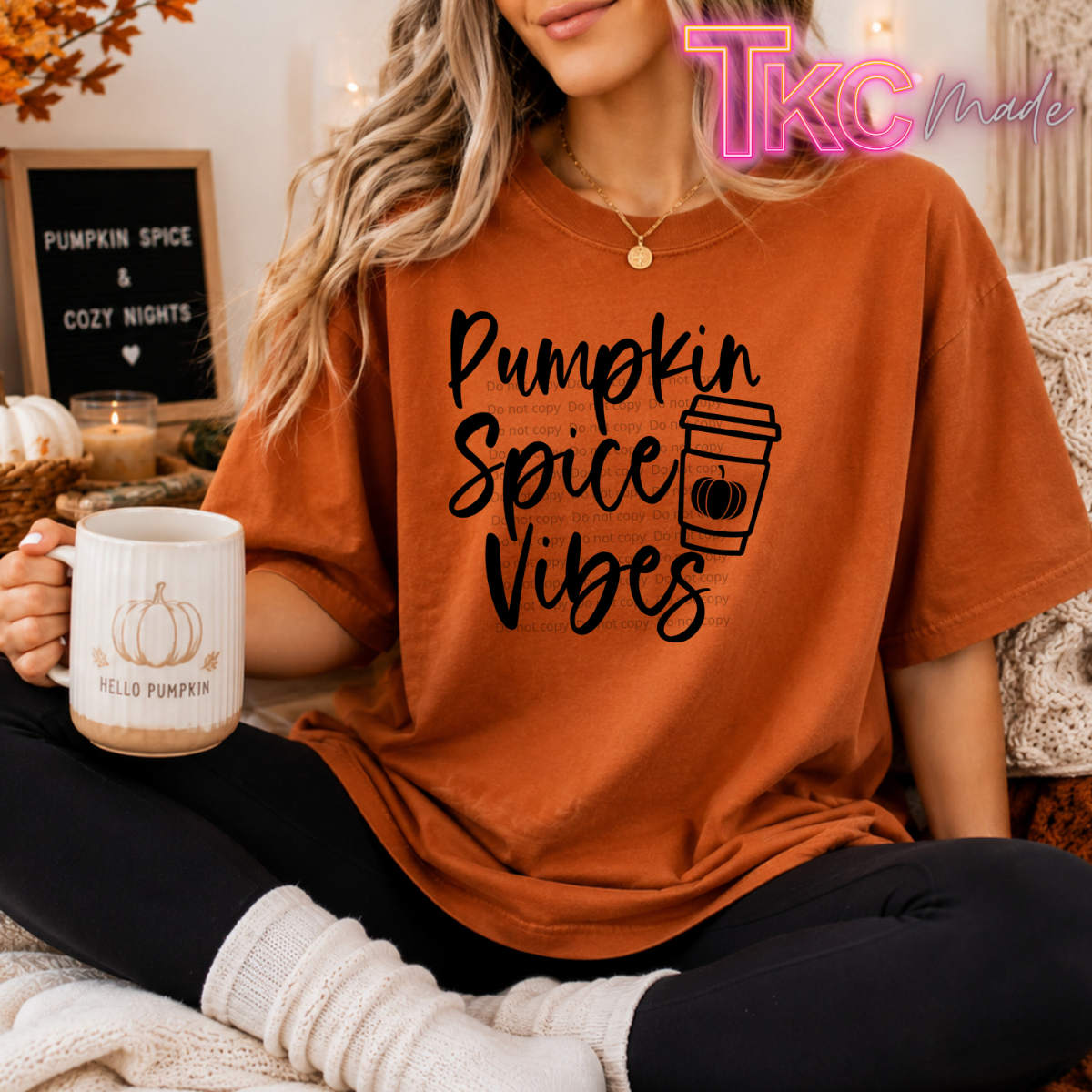 A woman wearing an oversized burnt orange graphic t-shirt with 'Pumpkin Spice Vibes' and a coffee cup illustration printed in black. She is sitting in a cozy room setting, holding a white mug with a pumpkin design and 'Hello Pumpkin' text.