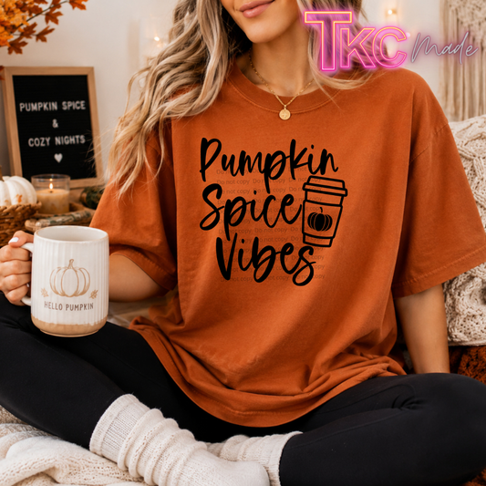 A woman wearing an oversized burnt orange graphic t-shirt with 'Pumpkin Spice Vibes' and a coffee cup illustration printed in black. She is sitting in a cozy room setting, holding a white mug with a pumpkin design and 'Hello Pumpkin' text.