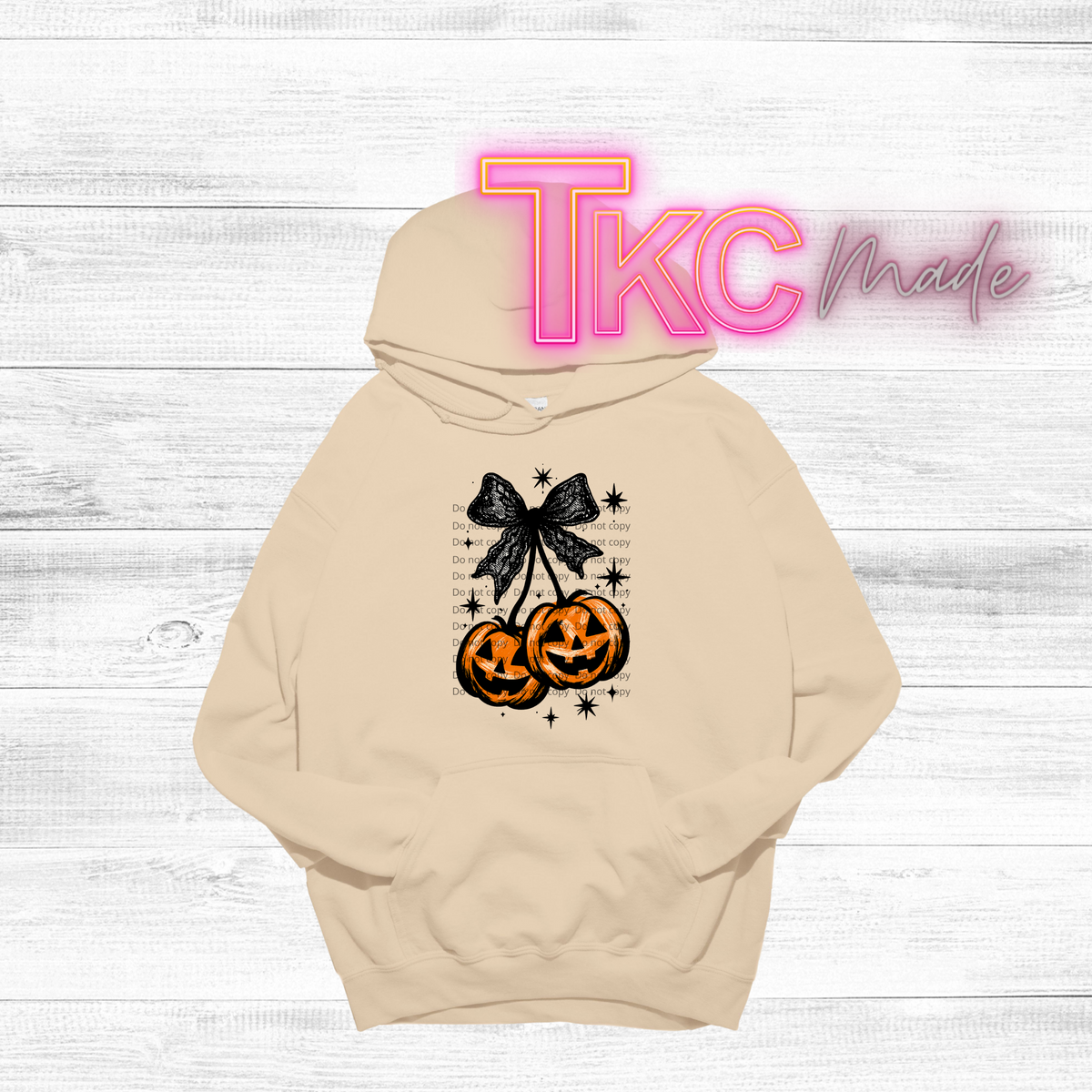A beige pullover hoodie with a Halloween graphic of three carved pumpkins, a black bow, and stars, displayed on a white wood background with a 'Tkc Made' watermark overlay.