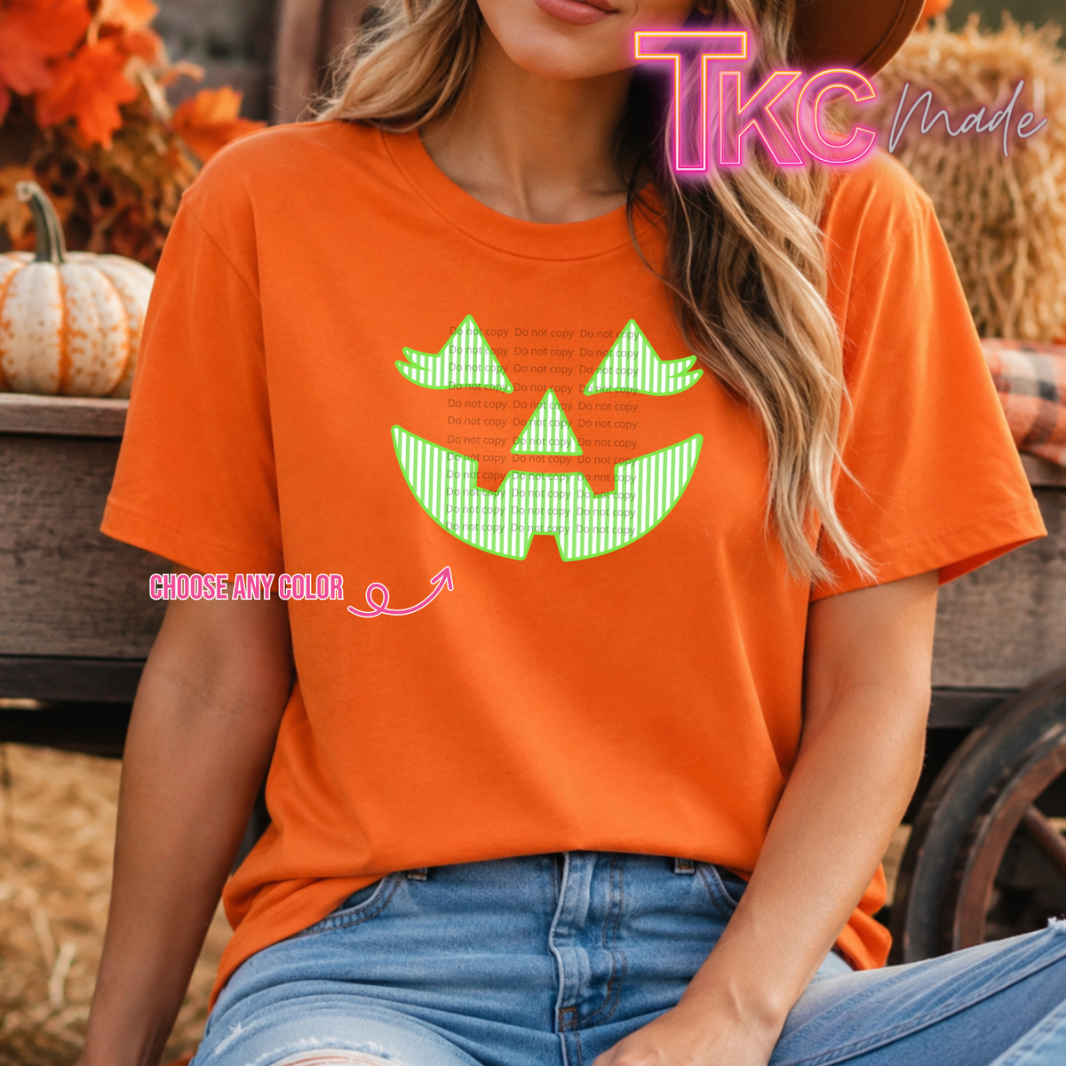 A woman wearing a bright orange short-sleeve T-shirt with a large green jack-o'-lantern face graphic on the front, paired with blue jeans in an outdoor autumn setting. The image includes 'TKC Made' branding and a 'CHOOSE ANY COLOR' 