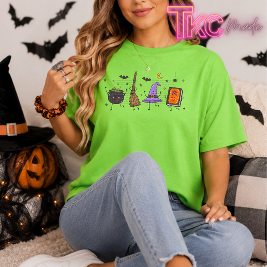 A woman wearing a bright lime green t-shirt with a colorful Halloween graphic of a cauldron, broom, witch hat, and potion bottle. She is sitting on a couch with Halloween decorations, including a jack-o'-lantern and black bat cutouts on the wall.
